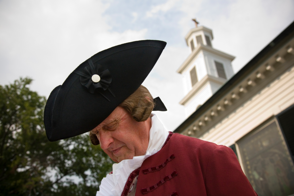 A colonial reenactor bows his head in front of a church